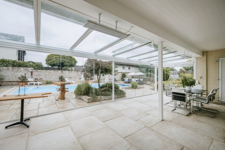 Terrassenverglasung COVER unter Glasdach ROOFS mit Blick auf Garten und Pool, rechts ein Essplatz