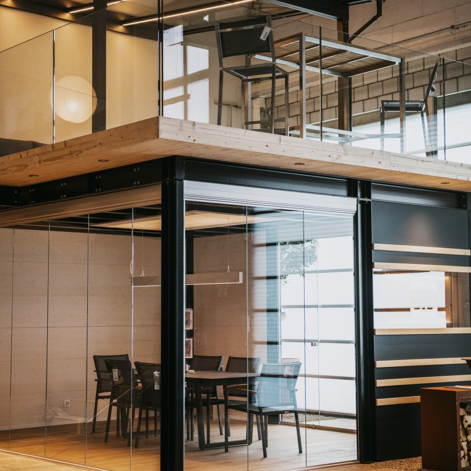 Seitenansicht des verglasten Besprechungsraums mit Tisch und Stühlen, darüber eine Galerie mit Glasbrüstung